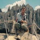 Ein lachender Mann in Wanderausrüstung kauert auf Felsen vor einer beeindruckenden Felsformation.