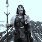 Eine Kriegerin steht mit Speer in einer verschneiten Berglandschaft und trägt Fellkleidung und Kriegsbemalung im Gesicht.