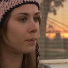 Eine Frau mit gestrickter Mütze blickt nachdenklich. Im Hintergrund ein Sonnenuntergang durch ein Fenster sichtbar.