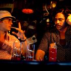 Zwei Männer in einem dunklen Raum, einer trägt Hut und Sonnenbrille, der andere hat eine Hand im Haar. Getränke auf dem Tresen.