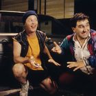 Zwei Männer knien lachend vor einem Auto. Der linke trägt Mütze und Weste, der rechte bunte Jacke. Beide halten Münzen.