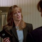 Eine Frau hält eine VHS-Kassette in der Hand und spricht mit einem Mann. Sie hat lange, blonde Haare und trägt einen Blazer.