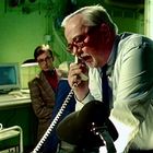 Zwei Männer in einem Büro, einer am Telefon, im Hintergrund ein alter Computer und Schreibtischlampen.