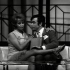 Es gibt keinen sichtbaren Text. Eine Frau sitzt neben einem Mann auf einem Sofa, sie schauen einander an. Schwarz-Weiß-Foto.