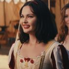 Zwei Frauen in mittelalterlichen Kostümen, die Hauptfrau lächelt, mit dunklem Haarband und weißen Kleid mit roten Details.