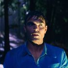 Ein Mann mit nassem Haar in einem blauen Hemd steht im dunklen Wald, umgeben von Regen und Bäumen.