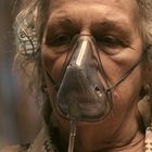 Eine ältere Person mit grauem Haar trägt eine Sauerstoffmaske und hat einen ernsten Gesichtsausdruck.