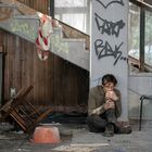 Ein junger Mann sitzt in einem verlassenen Raum, umgeben von Graffiti an Wänden und einer kaputten Treppe.