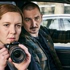 Ein Mann und eine Frau im Auto, die Frau hält eine Kamera und schaut fokussiert, der Mann schaut ernst zum Fenster hinaus.