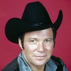 Es gibt keine sichtbaren Texte. Ein Mann trägt einen schwarzen Cowboyhut und schaut vor einem roten Hintergrund freundlich.
