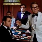 Männer in Anzügen an einem Spieltisch. Ein Mann in weißem Smoking steht auf und wirkt überrascht.