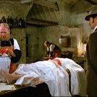 Text nicht sichtbar. Drei Männer in rustikalem Raum. Einer mit blutigen Händen, ein anderer in Anzug, im Hintergrund ein Tisch.