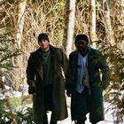 Zwei Männer in dicken Mänteln laufen durch einen schneebedeckten Wald. Die Sonne scheint durch die Bäume.