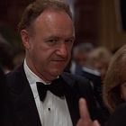 Es sind keine Texte zu sehen. Ein Mann in Anzug und Fliege steht in einer eleganten Umgebung mit mehreren Personen.