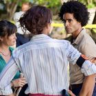 Person mit "POLICE" auf Armband diskutiert mit zwei anderen aufgeregt wirkenden Menschen im Freien, vor Absperrband.