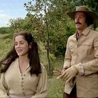 Eine Frau und ein Mann in historischer Kleidung stehen lächelnd in einer grünen, ländlichen Umgebung.
