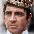 Ein Mann mit ernster Miene trägt eine verzierte Krone mit blauen und goldenen Edelsteinen.