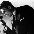 Ein Mann in einem Anzug spricht konzentriert in einen altmodischen Telefonhörer. Schwarz-Weiß-Aufnahme.