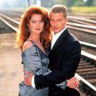 Ein Paar umarmt sich neben Bahngleisen. Die Frau hat rotes Haar, trägt ein glänzendes Kleid, der Mann einen dunklen Anzug.