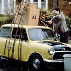 Auf einem gelben Auto ist ein großer Karton mit „TV“ beschriftet. Ein Mann befestigt ihn mit Seilen.