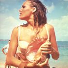 Eine Frau im Bikini hält eine große Muschel am Strand, mit einem bewölkten Himmel im Hintergrund.