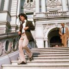 Zwei Männer gehen eine breite Steintreppe hinunter, vor einem prunkvollen Gebäude mit Säulen und Verzierungen.