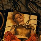 Eine Person mit blonden Haaren liegt auf einem Tisch, umgeben von Kabeln und goldenen Schläuchen, in einem goldenen Top.