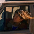 Junge Frau mit windzerzaustem Haar schaut nachdenklich aus einem Autofenster in einer sonnigen Umgebung.