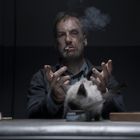 Ein Mann mit Verletzungen im Gesicht sitzt an einem Tisch, raucht eine Zigarette. Eine Katze schnuppert an einer Dose.