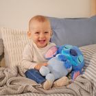 Ein lachendes Baby sitzt mit einem blauen Kuscheltier auf einem Bett mit grauen Decken und mehreren Kissen.