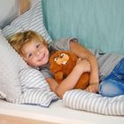 Ein Kind liegt lächelnd auf einem Bett, hält ein braunes Stofftier. Kissen und Decke sind gestreift.