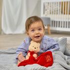 Ein lächelndes Baby sitzt auf einer grauen Decke, hält ein Kuscheltier und trägt ein blaues Hemd. Im Hintergrund ein weißes Gitterbett.