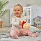 Ein fröhliches Baby sitzt auf einer Decke, hält einen kleinen Teddybär. Im Hintergrund Pflanzen und ein Babybett.