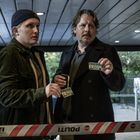 Zwei Männer halten Presseausweise hoch. Sie stehen hinter einem "POLITI" Absperrband in einem überdachten Bereich.