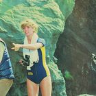 Auf einem Felsen stehen drei Personen: ein Mann mit Gewehr, eine Frau in Badeanzug mit Kamera, ein Mann mit Harpune.