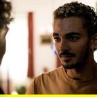 Eine Nahaufnahme von zwei Männern in einem Raum im Gespräch vertieft. Der rechte Mann hat lockiges Haar und trägt ein Ohrpiercing.