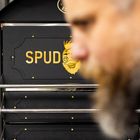 Der Text "SPUD" in Gelb ist auf einem schwarzen Hintergrund zu sehen. Im Vordergrund ist ein unscharfes Gesicht.