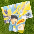 "SMILE" in roter Schrift, Maya Shleifer. Zwei Kinder schauen sich an, umgeben von hellen Farben. Buch auf grünem Gras.