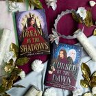 Zwei Bücher: "Dream by the Shadows" und "Cursed by the Dawn" von Logan Karlie. Reich an Blumen und goldenen Blättern dekoriert.
