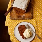 Ein Laib Kuchen mit Zuckerguss auf einem Holzbrett, daneben ein Messer. Ein Teller mit einem Kuchenstück und Sahne.