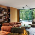 Ein Wohnzimmer mit großen Fenstern, modernen Sofas in Brauntönen, einem Glastisch und einem Bücherregal. Blick auf Bäume.