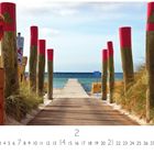 "Weg zum Meer" über einem hölzernen Steg, flankiert von rot markierten Pfählen, führt zum Strand mit blauen Himmel.