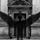 Zwei Fenster mit Gittern flankieren eine dekorative Tür. Ein Schatten in Form von Flügeln erstreckt sich über die Fassade.