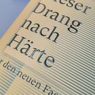„Dieser Drang nach Härte“ steht in großen Buchstaben auf einem hellen Hintergrund mit dunklen, schmalen Streifen.