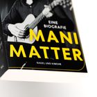 Text: "EINE BIOGRAFIE MANI MATTER NAGEL UND KIMCHE". Schwarz-Weiß-Foto eines Mannes mit Gitarre.