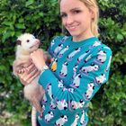 Keine Texte vorhanden. Eine Frau in einem bunten Pullover mit Tiermotiven hält ein Opossum. Hintergrund ist grün bewachsen.