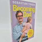 "SEBASTIAN TIGGES Becoming Dad" steht auf dem Cover eines Buches. Ein Mann hält ein Kind, lachend vor hellem Hintergrund.