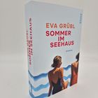 "Eva Grübl - Sommer im Seehaus - Roman." Zwei Frauen in Badeanzügen sitzen am Wasser; ein blauer Wellenhintergrund.