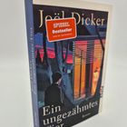"Ein ungezähmtes Tier" von Joël Dicker. SPIEGEL Bestseller-Aufkleber. Ein Mann schaut auf ein beleuchtetes Fenster.