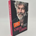 Text: "Spiegel Bestseller", "ÜBER LEBEN", "Reinhold Messner". Ein Stapel Bücher mit einem Porträt eines Mannes vorne drauf.